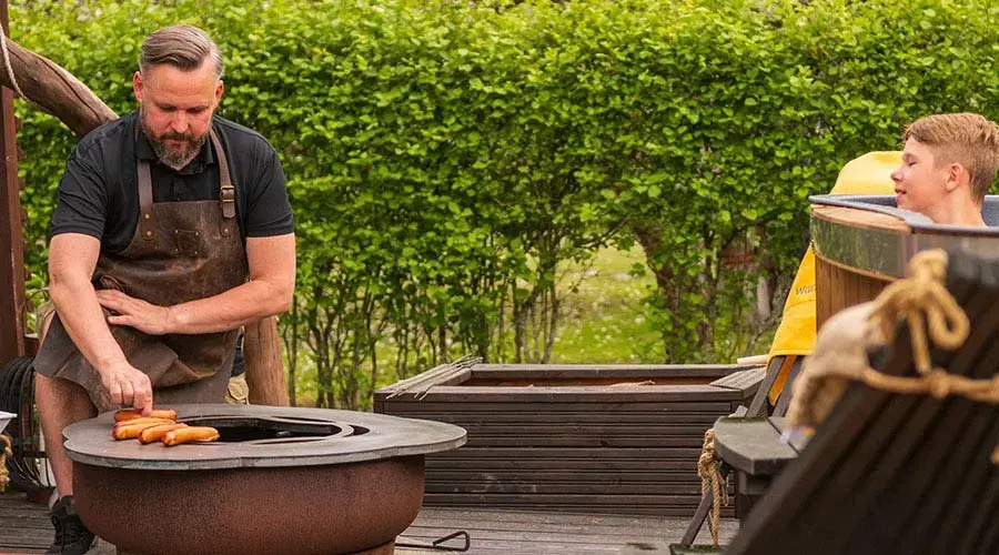 Man in schort bakt vlees op een grill, terwijl een jongen toekijkt in een tuin.